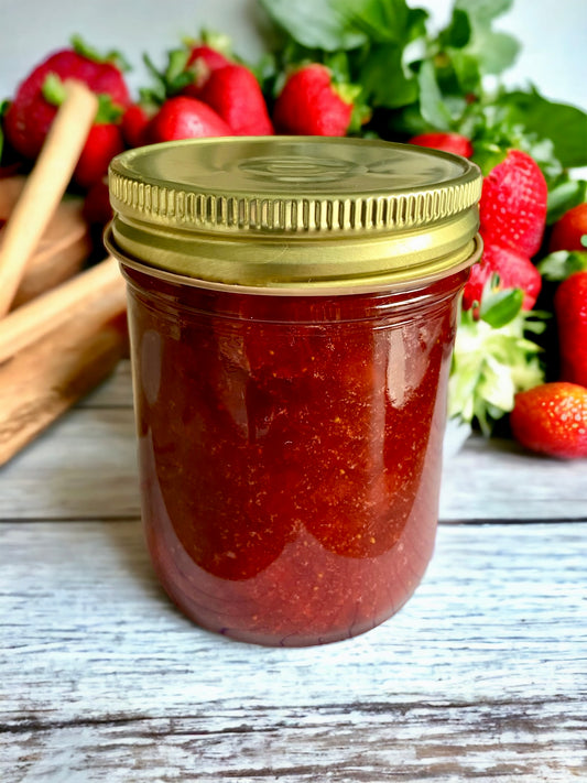 A jar of strawberry rhubarb jam on a rustic table with strawberries and rhubarb in the background.