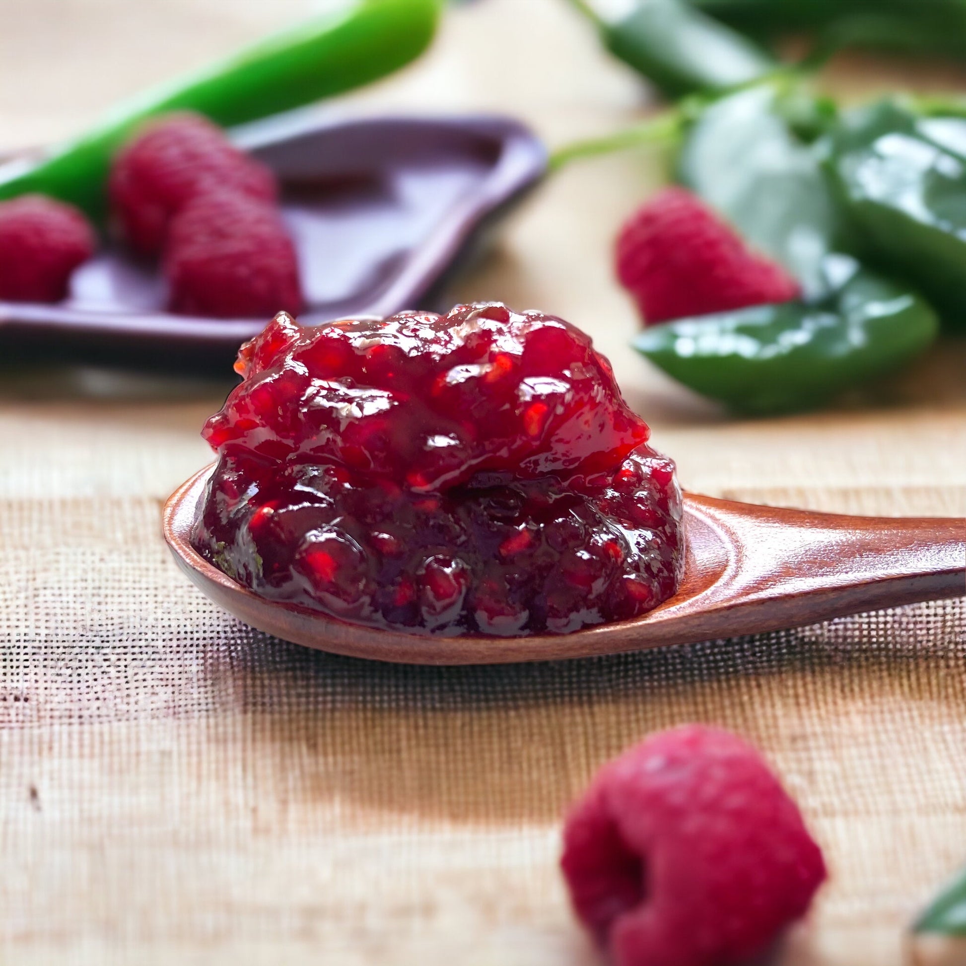 a wooden spoon loaded with raspberry jalapeño jam resting on a linen cloth with fresh raspberries and jalapeno peppers all around